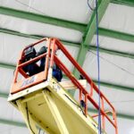 Yellow and red scissor lift raised close to the steel roof trusses of an industrial building